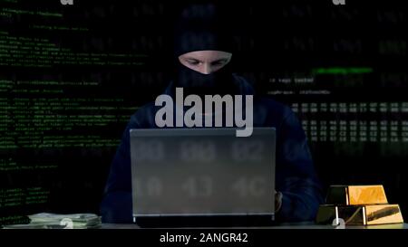 Criminal in balaclava working on laptop, hacking bank security system, data code Stock Photo