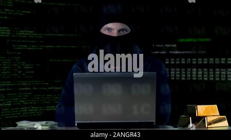 Hacker in balaclava committing cyber crime on laptop, numbers codes background Stock Photo