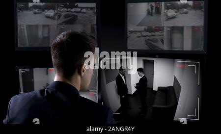 Male security operator watching surveillance cameras, illegal business deal Stock Photo