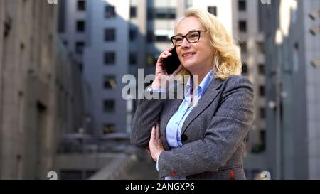 Cheerful female manager talking on phone standing outside, client communication Stock Photo