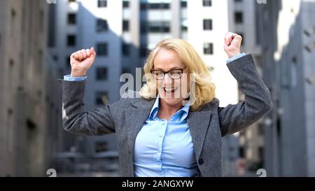 Excited mature female boss in formal suit dancing outdoors, work motivation Stock Photo