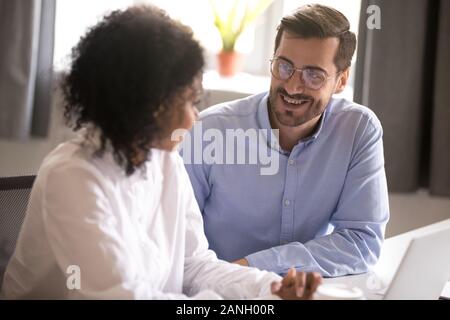 Smiling man talk with female colleague working together on project at laptop, diverse millennial coworkers laugh chatting or having conversation while Stock Photo