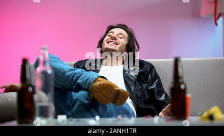 Relaxed drunk young man sitting on sofa, boozer enjoying music at dance party Stock Photo