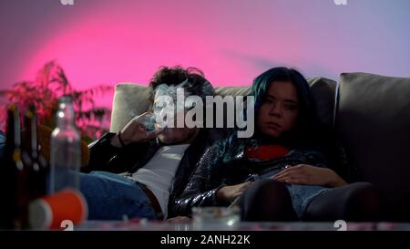 Drunk couple of druggies sitting on coach at home party in messy room, bad habit Stock Photo