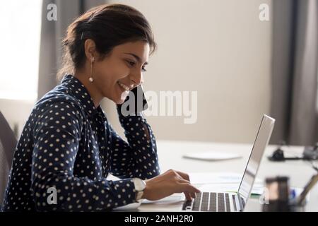 Smiling female indian employee multitask working on computer in office talking over phone, happy woman worker using laptop, laughing speaking with fri Stock Photo