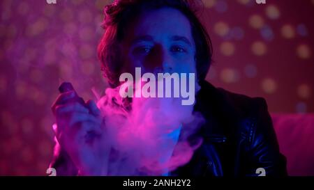 Thoughtful unhappy man exhaling smoke of e-cigarette in nightclub lights, vaping Stock Photo