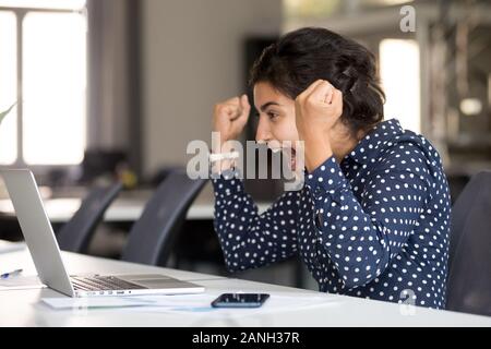 Excited millennial employee sit at desk witness online lottery win on laptop, scream from happiness, happy indian woman winner look at computer screen Stock Photo