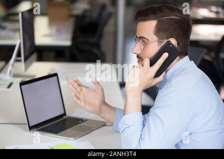 Male employee sit at office desk consulting client or customer on phone, serious businessman talk on smartphone with business partner or colleague, mi Stock Photo