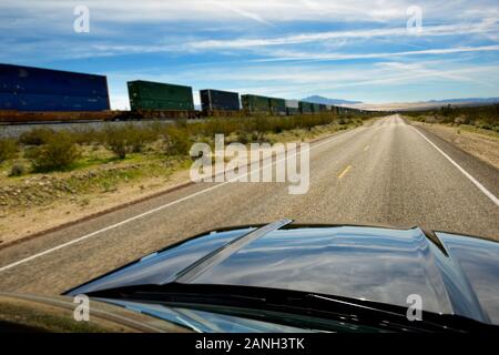 Car driving on deserted, straight freeway portion, parallel to railroad with long cargo train. Stock Photo