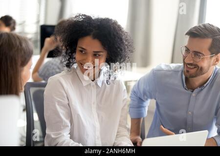Smiling multiracial workers talk chatting while working together on project on laptop, diverse employees brainstorm speaking at workplace discussing i Stock Photo