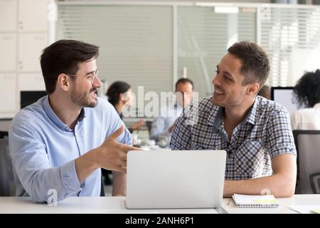 Smiling male employees work together on laptop in shared office, men cooperating using computer discussing business ideas on workplace, excited worker Stock Photo