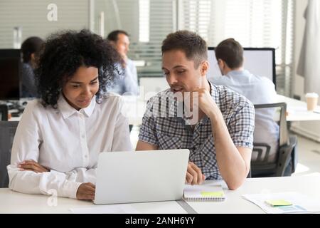 Multiracial millennial colleagues sit at office desk working on laptop discussing project or startup, focused diverse employee brainstorm watching tra Stock Photo