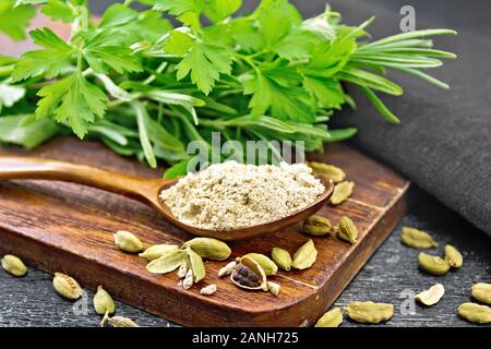 Ground cardamom in a spoon, seasoning capsules, napkin, fresh parsley and rosemary on black wooden board background Stock Photo