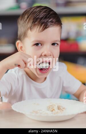 Cute baby eats milk porridge. Beautiful boy has breakfast sitting at a table in the kitchen at home. Stock Photo