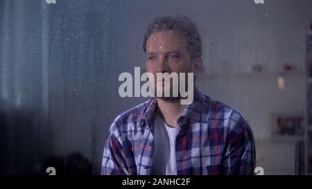 Lonely sad middle-aged man looking through rainy window, bad weather conditions Stock Photo
