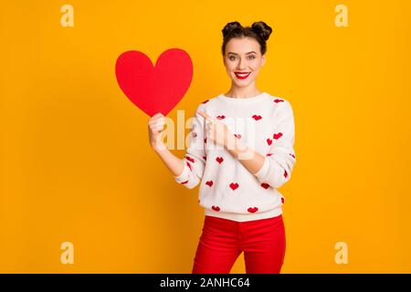 Photo of pretty affectionate lady hold paper heart show date postcard indicating finger promotion wear white red hearts pattern pullover pants Stock Photo