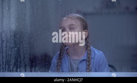 Sad refugee girl looking through rainy window in rehabilitation center, recovery Stock Photo