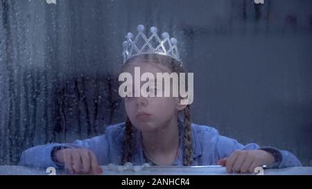 Upset little girl in crown with magic stick sitting alone behind rainy window Stock Photo