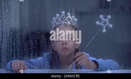 Lonely little princess in crown with magic stick sitting behind rainy window Stock Photo