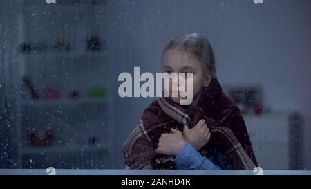 Sick girl covered in blanket looking through rainy window, rehabilitation center Stock Photo