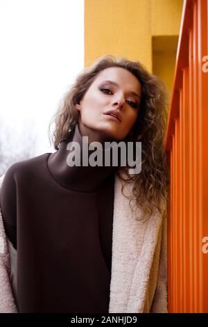 A girl with red curly hair in a white coat poses on the yellow orange parking stairs. City Style - Urban Stock Photo