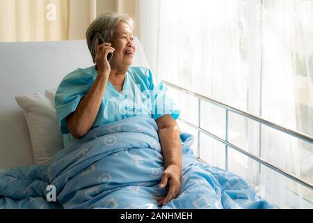 Asian senior elderly using chatting to family on her mobile phone and smiling, with delight as she listens to the conversation while she illness in ho Stock Photo