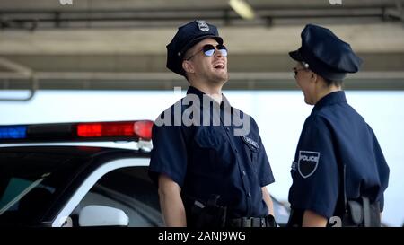 Young police officers laughing standing near car, successful patrol shift Stock Photo