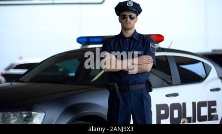 Brave police officer in service cap and sunglasses posing into camera near car Stock Photo