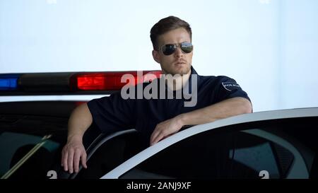 Serious patrol officer in sunglasses monitoring road standing near car door Stock Photo