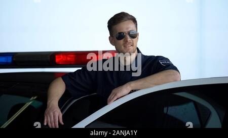 Friendly policeman in sunglasses standing near car and smiling, law and order Stock Photo