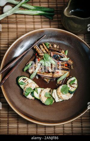 Spicy Aubergine / Eggplant + tofu & vegetable stir fry, served on a ceramic plate,  chopsticks, Bamboo matt & steamer. Black background. Flat lay Stock Photo