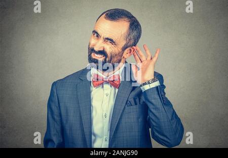 A mature, middle-aged man holds his hand near ear and listening trying to hear. hearing, deaf problem concept. Mixed race bearded model isolated on gr Stock Photo