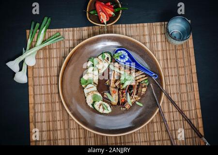 Spicy Aubergine / Eggplant with tofu & vegetable stir fry, served on a plate with chopsticks. Bamboo matt & steamer with black background. Flat lay Stock Photo