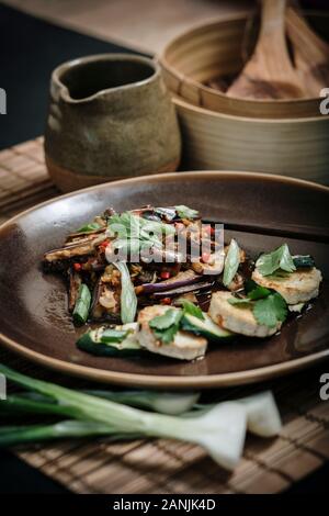 Vegan recipe of Asian spicy aubergine with garlic, tofu, chilli and spring onions served on a plate with a bamboo matt and chopsticks Stock Photo