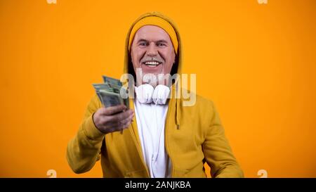 Happy old man in earphones and stylish clothes holding dollars and having fun Stock Photo