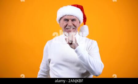 Happy gray bearded man in Santa Claus hat showing silence gesture and smiling Stock Photo