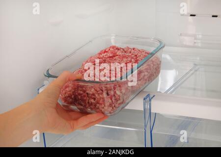 Minced raw meat in glass container is on shelf in open fridge. Female hand pulls out from fridge an ingredient for cooking meat dishes. Stock Photo
