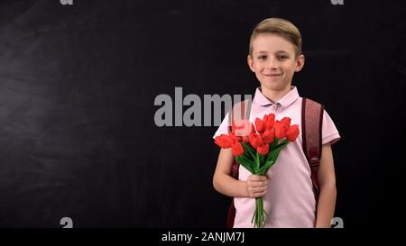 Boy holding tulips standing near chalkboard, congratulating teacher, first love Stock Photo