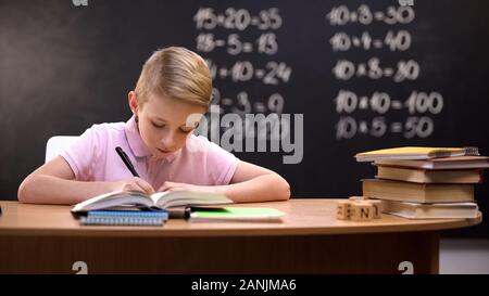 Schoolboy doing sums on math lesson, sitting against blackboard, knowledge Stock Photo