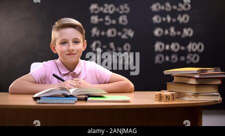 Smart schoolboy smiling to camera after solving task, exercises written behind Stock Photo