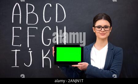 Smiling teacher holding prekeyed tablet, alphabet on blackboard, innovations Stock Photo