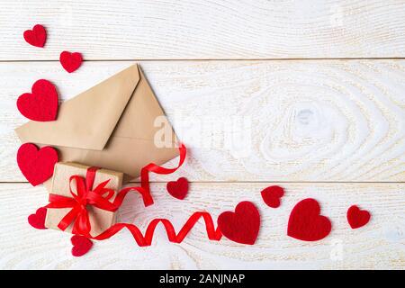 Gift box with red ribbon bow, brown paper envelope and valentine hearts on a white wood textured surface. Greeting card with copy space. Love concept. Stock Photo