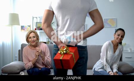 Curious wife and mother-in-law looking at man holding gift boxes behind back Stock Photo