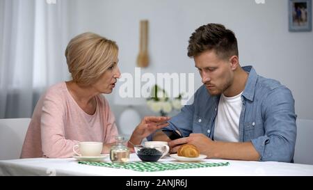 Worried mother talking to infantile adult son playing video games on smartphone Stock Photo
