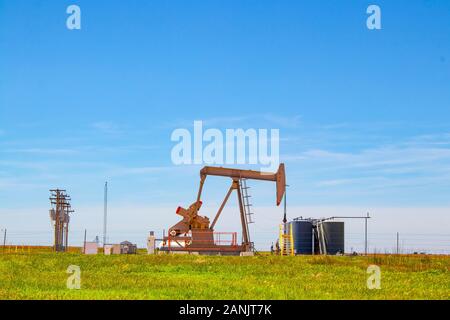 Working Pump Jack on oil well with  tanks on site out on the horizon on the plains with electric lines and blue sky in background Stock Photo