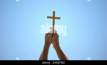 Male hands raising wooden cross to blue sky, religious conversion, baptism Stock Photo