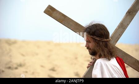 Jesus in crown of thorns carrying heavy cross, sacrifice for sinners salvation Stock Photo