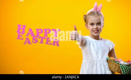 Happy Easter text, cute child with eggs basket in bunny ears showing thumbs up Stock Photo