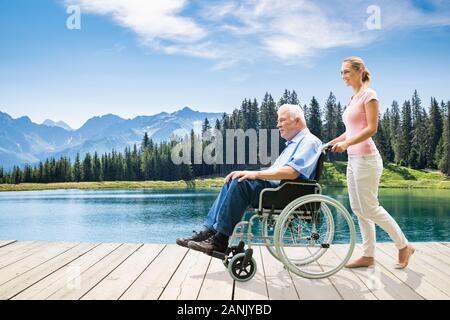 Young Woman Assisting Her Disable Grandfather In Wheelchair Walking On Boardwalk Near Lake Stock Photo