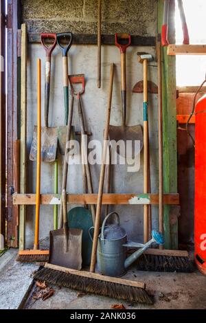Old gardening tools including spades, brushes and watering can stacked against an old shed wall Stock Photo